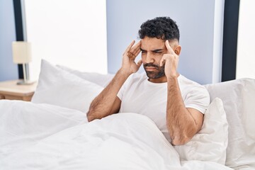 Young hispanic man suffering for headache sitting on bed at bedroom