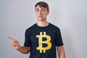 Caucasian blond man wearing bitcoin t shirt pointing aside worried and nervous with forefinger,...