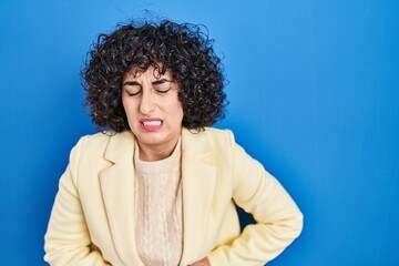 Young brunette woman with curly hair standing over blue background with hand on stomach because indigestion, painful illness feeling unwell. ache concept.