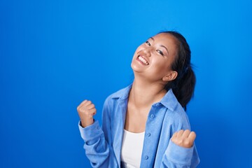 Fototapeta premium Asian young woman standing over blue background very happy and excited doing winner gesture with arms raised, smiling and screaming for success. celebration concept.