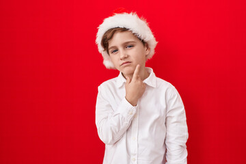 Little caucasian boy wearing christmas hat over red background thinking concentrated about doubt...