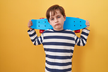 Little caucasian boy holding skate over yellow background relaxed with serious expression on face....