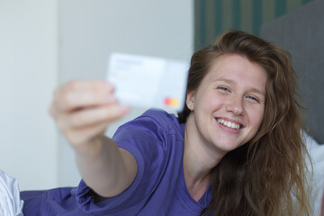 Young happy woman doing online shopping with smartphone, lay in bed in bedroom at home, hold in hand credit bank card and cell mobile phone and smile