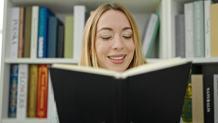Young blonde woman student smiling confident reading book at library university