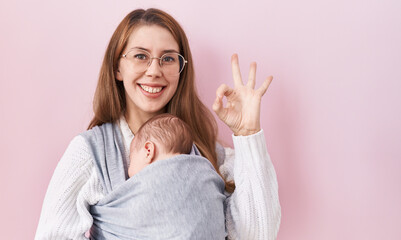 Young caucasian woman holding and carrying baby on a sling doing ok sign with fingers, smiling...