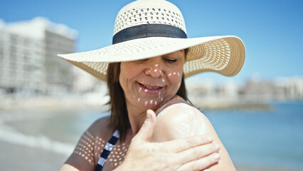 Middle age hispanic woman tourist smiling wearing summer hat applying sunscreen at the beach