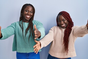 Two african woman standing over blue background looking at the camera smiling with open arms for...