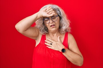Middle age woman with grey hair standing over red background touching forehead for illness and fever, flu and cold, virus sick