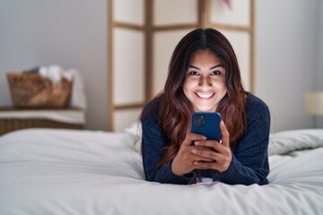 Young hispanic woman using smartphone lying on bed at bedroom