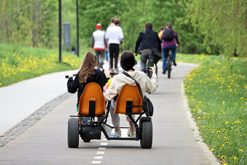 Two girls riding velomobile in a green park. Tandem bike renting for spring or summer leisure