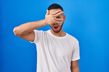 Young hispanic man standing over blue background peeking in shock covering face and eyes with hand,...