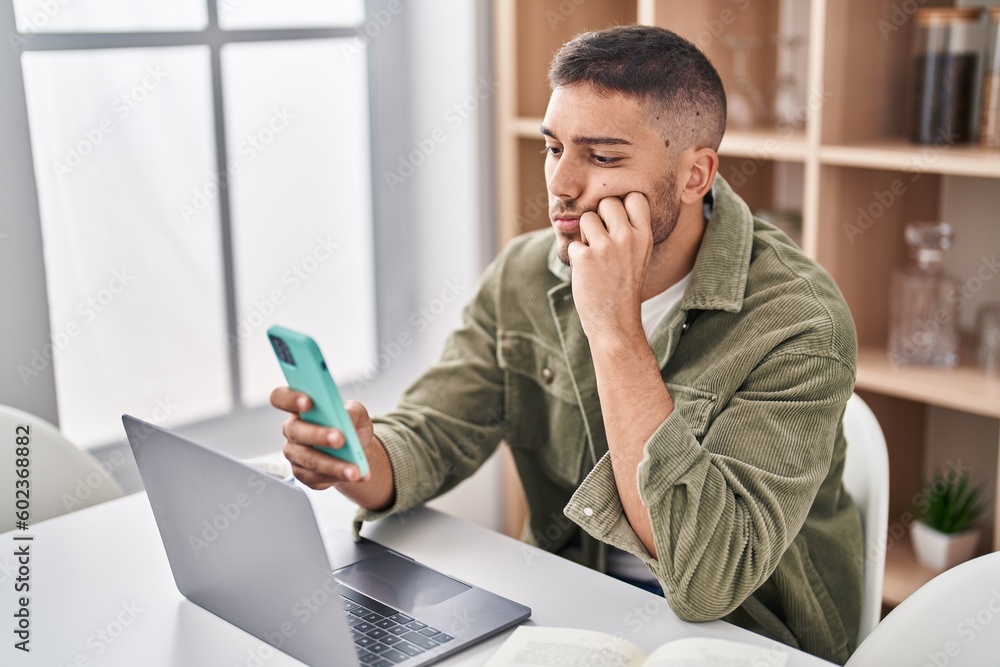 Wall mural Young hispanic man sitting on table using smartphone at home