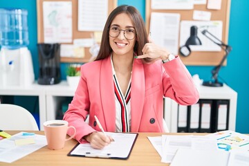 Young hispanic woman working at the office wearing glasses smiling doing phone gesture with hand and fingers like talking on the telephone. communicating concepts.