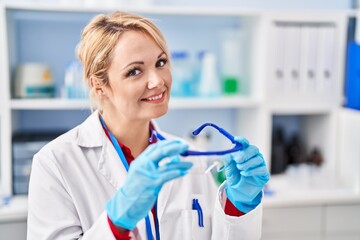 Young blonde woman scientist smiling confident holding glasses at laboratory