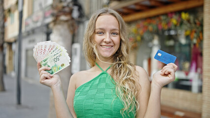 Young blonde woman holding credit card and israel shekels at street