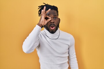 African man with dreadlocks wearing turtleneck sweater over yellow background doing ok gesture shocked with surprised face, eye looking through fingers. unbelieving expression.