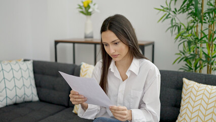 Fototapeta premium Young beautiful hispanic woman reading document sitting on sofa at home