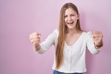 Young caucasian woman standing over pink background angry and mad raising fists frustrated and...