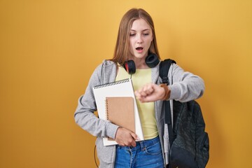 Young caucasian woman wearing student backpack and holding books looking at the watch time worried, afraid of getting late