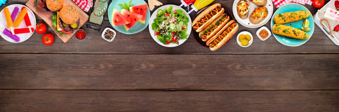 Summer BBQ food top border. Hamburgers, hot dogs, potatoes, corn and cold treats. Overhead view over a dark wood banner background. Copy space.