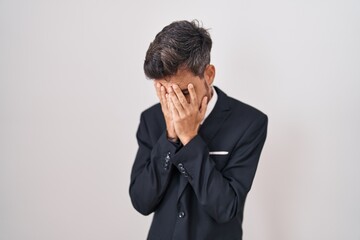 Young hispanic man with tattoos wearing business suit and tie with sad expression covering face...