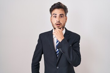 Young hispanic man with tattoos wearing business suit and tie looking fascinated with disbelief, surprise and amazed expression with hands on chin