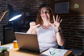 Brunette woman working at the office at night showing and pointing up with fingers number six while smiling confident and happy.