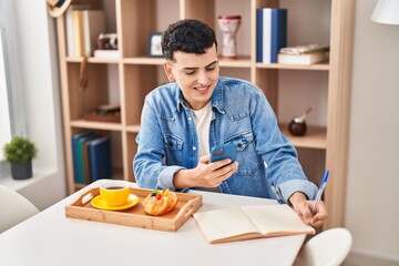 Young non binary man using smartphone and writing on notebook having breakfast at home