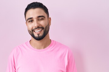 Hispanic young man standing over pink background winking looking at the camera with sexy expression, cheerful and happy face.