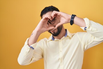 Hispanic young man wearing business clothes and glasses doing heart shape with hand and fingers smiling looking through sign