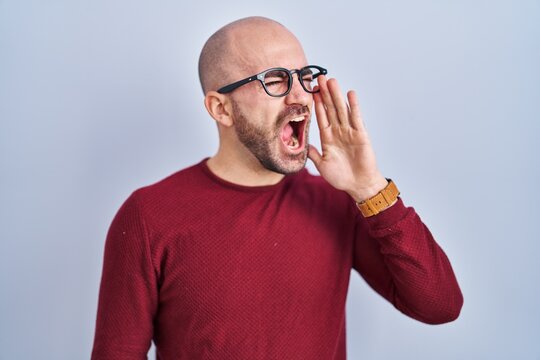 Young bald man with beard standing over white background wearing glasses shouting and screaming loud to side with hand on mouth. communication concept.