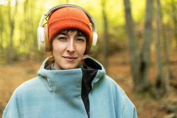short-haired woman in the forest with headphones smiling on green background, listening to music looking at the camera with a wool hat