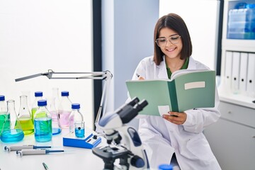 Young beautiful hispanic woman scientist smiling confident writing notebook at laboratory