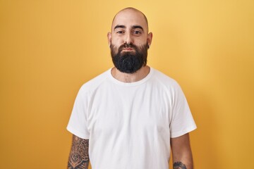 Young hispanic man with beard and tattoos standing over yellow background puffing cheeks with funny face. mouth inflated with air, crazy expression.