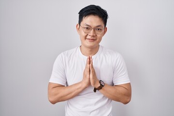 Young asian man standing over white background praying with hands together asking for forgiveness smiling confident.