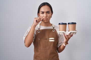 Young hispanic woman wearing professional waitress apron holding coffee mouth and lips shut as zip...