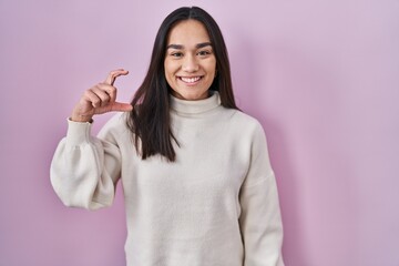 Young south asian woman standing over pink background smiling and confident gesturing with hand doing small size sign with fingers looking and the camera. measure concept.