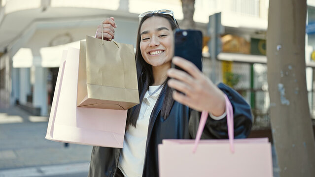 Young beautiful hispanic woman make selfie by smartphone holding shopping bags at street