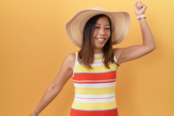 Middle age chinese woman wearing summer hat over yellow background dancing happy and cheerful, smiling moving casual and confident listening to music