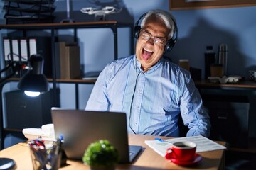 Hispanic senior man wearing call center agent headset at night sticking tongue out happy with funny expression. emotion concept.