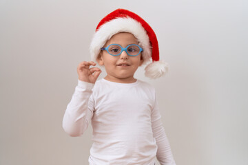 Little hispanic boy wearing glasses and christmas hat doing ok sign with fingers, smiling friendly gesturing excellent symbol