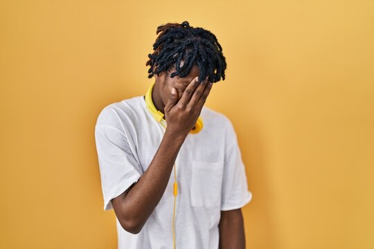Young african man with dreadlocks standing over yellow background with sad expression covering face with hands while crying. depression concept.