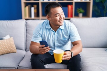 Young chinese man using smartphone drinking coffee at home