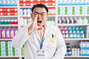 Chinese young man working at pharmacy drugstore shouting and screaming loud to side with hand on...