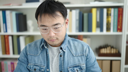 Young chinese man student sitting on table with relaxed expression at library university