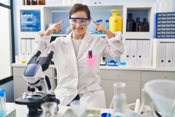 Hispanic girl with down syndrome working at scientist laboratory smiling pointing to head with both hands finger, great idea or thought, good memory