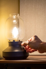 Male hand turns on vintage lantern lamp switch on wooden table in the dark room. Lighting equipment...