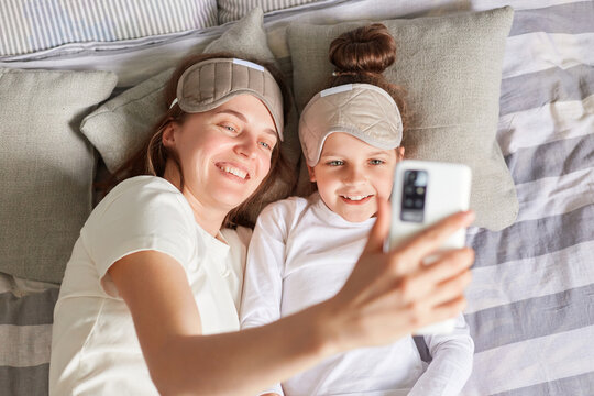 Top view of cheerful joyful mother and daughter waking up in morning in sleeping masks taking selfie on smart phone smiling to mobile phone camera.
