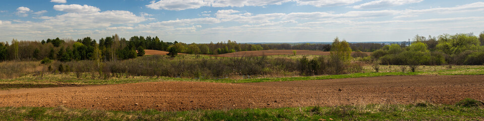spring agricultural landscape. plowed hilly fields with thickets under a blue cloudy sky. widescreen panoramic side view
