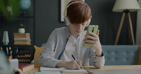 Teenage boy learning online listening to lecture through headphones and looking at smartphone screen and writing information at home. Modern technology and distant education concept.